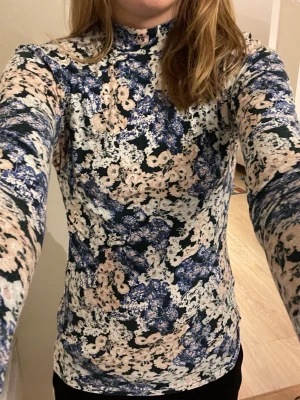 Blommig långärmad polotopp st S - Snygg långärmad polotopp från Carin Wester med blommigt mönster i blått, beige och vitt. Toppens material känns stretchigt och sitter tight mot kroppen men är också väldigt mjukt. Storlek S.