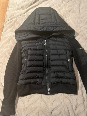 Svart montörer cardigan  - Snygg svart moncler cardigan med quiltad front och stor huva. Jackan har stickade ärmar och muddar, två fickor med tryckknappar och dragkedja framtill. Perfekt för kalla dagar 