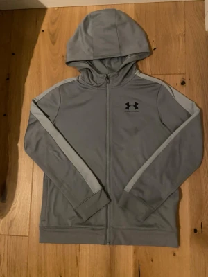 Grå hoodie från Under Armour - Snygg grå hoodie från Under Armour med dragkedja och huva. Tröjan har svarta loggan tryckt på bröstet och ljusare grå detaljer längs ärmarna. Tillverkad i ett mjukt och stretchigt material som känns sportigt och bekvämt.