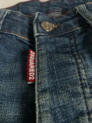 Blå jeans från Dsquared2 - Säljer ett par klassiska blå jeans från Dsquared2 med snygga slitningar och kontrastsömmar. Jeansen har en metallknapp med logotyp och en röd tygpatch med Dsquared2 på framsidan. Materialet är denim och passformen är rak.