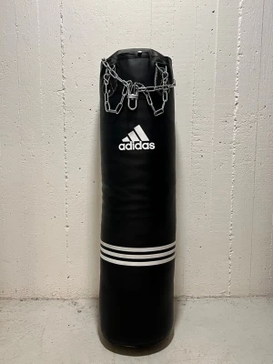 Boxningssäck från Adidas med kedja - Svart boxningssäck från Adidas med klassisk vit logga och kraftig metallkedja for upphängning. Tillverkad i slitstarkt konstläder som gör den perfekt för slag-och sparkträning. Höjd x bredd - 120cm x 33cm. Vikt ca 35kg. Passar både nybörjare och erfarna fighters inom kampsport, boxning. Använd bara ett par gånger, legat i förvaring sedan dess. Välkommen att hämtas upp i Handen, Stockholm. Transport går också att ordna i Stockholm för en kostnad av 250:-. Testar lägga ut här. 