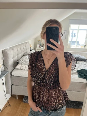 Leopardmönstrad blus med volang - Snygg blus med leopardmönster och volangdetaljer. Blusen har en djup v-ringning och korta, transparenta ärmar. Materialet är lätt och luftig, perfekt för att skapa en trendig look. Passar till jeans eller kjol för en cool stil.