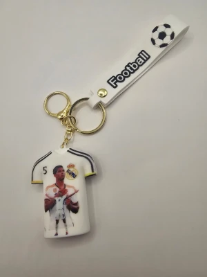 Nyckelring Real Madrid Bellingham - Cool nyckelring med vit Real Madrid-tröja, nummer 5 och Bellingham på baksidan. Tröjan har klubbens logga och detaljer i svart och gult. Nyckelringen har även ett vitt band med texten 'Football' och en fotbollssymbol. Perfekt för fotbollsfans!