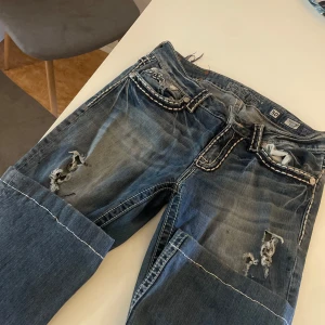 Miss Me bootcut jeans med slitningar - Säljer ett par blå Miss Me jeans i bootcut-modell med coola slitningar och kontrastsömmar. Jeansen har låg midja och detaljerade fickor med broderade kanter. Pris kan diskuteras, waist 30 men passar mer w28 och 29