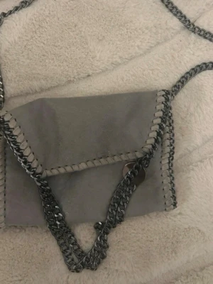 Grå stella mcartney liknande axelväska med kedjedetaljer - Grå axelväska i mocka med silverfärgade kedjor längs kanterna och som axelrem. Väskan har ett lock och dekorativ söm runt hela väskan. Perfekt för dig som gillar en stilren men edgy look.