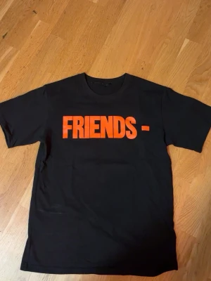 Svart Friends- t-shirt från VLONE - Cool svart t-shirt från VLONE med stor orange 'FRIENDS-' text på framsidan och ett stort orange V på ryggen. Klassisk rund hals och korta ärmar. Perfekt för dig som gillar streetwear och vill sticka ut med en statement-tee.