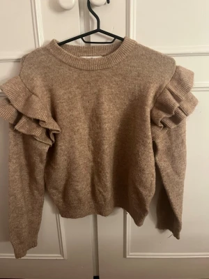 Beige stickad tröja med volangärmar - Mysig beige stickad tröja med rund halsringning och unika volangdetaljer på axlarna. Tröjan har långa ärmar och ribbstickade muddar vid hals, ärmslut och nederkant. Perfekt för dig som vill ha en trendig och bekväm look.