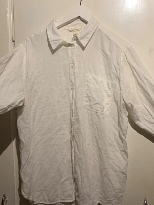 Vit skjorta i bomull med ficka - Klassisk vit skjorta i bomull med krage, bröstficka och knappar framtill. Skjortan har lång ärm och en avslappnad passform, perfekt för en clean och enkel stil. Passar till många olika outfits och är ett måste i garderoben.