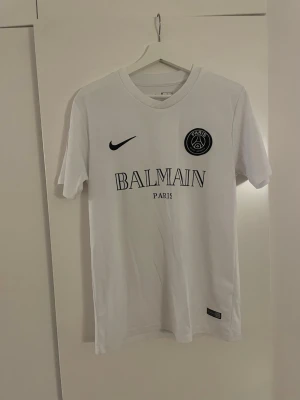 Vit PSG x Balmain t-shirt - Snygg vit t-shirt med Paris Saint-Germain-logga och Balmain Paris-tryck på bröstet. Tröjan har rund halsringning och är tillverkad i ett lätt och bekvämt material, perfekt för sport eller streetwear. Nike-logga på bröstet ger extra stilpoäng.