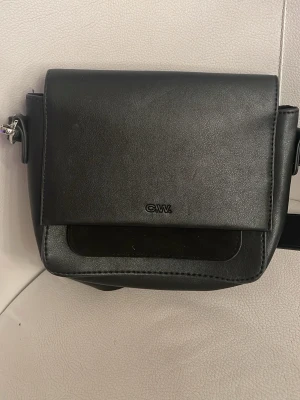 Svart axelväska från G.W. - Stilren svart axelväska från G.W. i slätt konstläder med justerbar bred axelrem och lockstängning. Väskan har minimalistisk design med diskret logga framtill och silvriga metalldetaljer. Perfekt för att bära det viktigaste med stil.