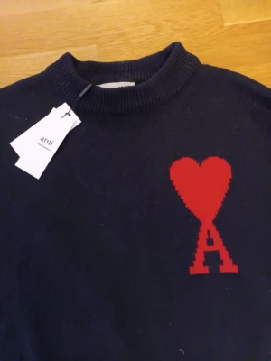 Svart stickad tröja från Ami Paris - Snygg svart stickad tröja från Ami Paris med ikoniskt rött hjärta och A-logga på bröstet. Tröjan har ribbad rund hals och är tillverkad i mjukt material som känns lyxigt. Perfekt för dig som gillar stilrena detaljer och streetwear-vibe.