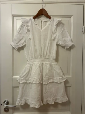 Sommarklänning- vit med volanger och kort ärm - Söt vit klänning med volanger och korta ärmar. Klänningen har v-ringning, resår i midjan och dubbla volanglager på kjolen. Materialet känns som bomull eller linne och ger en luftig känsla. Perfekt för varma dagar och har fina detaljer vid axlar och ärmslut.