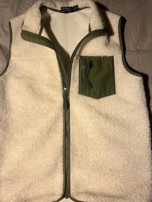 Fleeceväst från Polo Ralph Lauren - Säljer en krämvit fleeceväst från Polo Ralph Lauren med gröna detaljer och dragkedja framtill. Västen är superskön samt varm så perfekt nu inför hösten. Den är också nytvättad och fräsch. Säljs nu för ett sjukt bra pris!