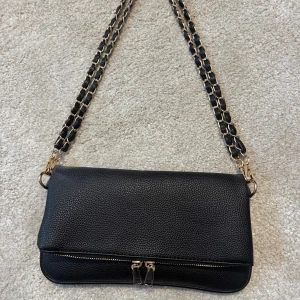 Svart handväska med kedjerem - Snygg svart handväska i konstläder med struktur och guldfärgade detaljer. Väskan har en dubbel kedjerem med invävt svart band och dragkedja framtill samt ett extra fack med dragkedja på baksidan. Perfekt för dig som gillar stilrena accessoarer.