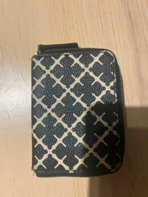 Svart mönstrad plånbok By Malene Birger - Snygg svart plånbok från By Malene Birger med beige grafiskt mönster och dragkedja runtom. Insidan är ljusrosa med plats för kort och kontanter. Perfekt storlek för fickan eller väskan och tillverkad i tåligt material.