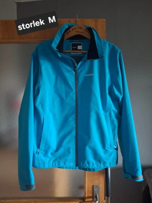 Blå vindjacka från FCURO - Snygg blå vindjacka från FCURO med dragkedja framtill och två sidofickor med dragkedja. Jackan har en hög krage och diskret logga på bröstet. Perfekt för blåsiga dagar och enkel att matcha med olika outfits. Använd en gång