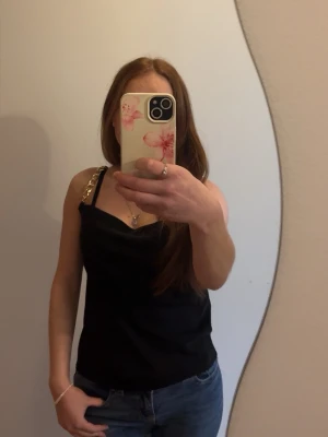 Svart linnetopp med gulddetaljer - Snygg svart linnetopp med smala axelband och guldfärgade paljetter på ena axelbandet. Toppen har en enkel och stilren design med lätt draperad front. Storlek XS