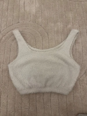Vit fluffig loungewear topp - Supermjuk och fluffig vit loungewear topp med breda axelband och rundad halsringning. Perfekt för mysiga hemmadagar och chillkvällar. Tillverkad i ett gosigt, teddy-liknande material som känns riktigt skönt mot huden.