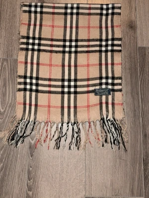 Beige rutig halsduk från Burberrys - Klassisk beige halsduk från Burberrys i 100% kashmir med det ikoniska rutmönstret i svart, vitt och rött. Halsduken har fransar i båda ändar och en vävd etikett. Perfekt accessoar för att lyfta din outfit.
