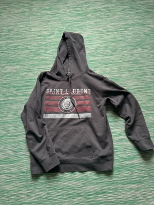 Mörkgrå Saint Laurent hoodie - Snygg mörkgrå hoodie från Saint Laurent med tryck framtill i vitt och rött. Klassisk huva med dragsko och stor magficka. Perfekt för en avslappnad streetwear-look. Materialet känns mjukt och bekvämt, troligen bomullsblandning.passar dig runt 165-175 cm