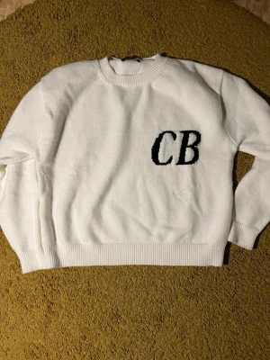 Vit stickad tröja CB Ole Brumm - Säljer en vit stickad tröja från OLE BRUMM med svarta bokstäver 'CB' broderat på bröstet. Tröjan har rund halsringning, ribbade muddar och är mjuk i materialet. Perfekt för dig som gillar enkel men snygg design.