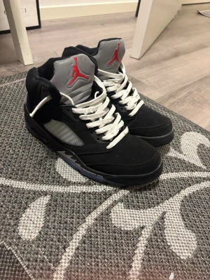 Svarta Air Jordan 5 Retro sneakers - Snygga Air Jordan 5 Retro sneakers i svart mocka med vita snören och grå plös med röd Jumpman-logga. Skorna har genomskinliga detaljer på sidorna och en robust sula med isblå inslag. Perfekta för dig som gillar klassisk streetwear-stil.