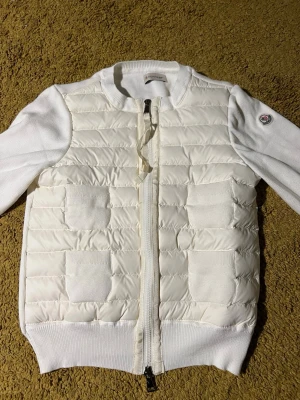 Vit damcardigan från Moncler - Snygg vit tröja från Moncler med quiltad front och stickade ärmar. Tröjan har dragkedja framtill, två fickor och ribbade muddar. Moncler-logga på ärmen ger en exklusiv touch. Perfekt för dig som gillar stilrena och sportiga plagg. Allt medföljer KVITTO,dustbag,etiketter,