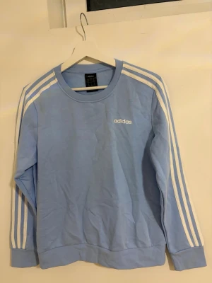 Ljusblå Adidas sweatshirt med ränder - Ljusblå sweatshirt från Adidas med klassiska vita ränder längs ärmarna och liten Adidas-logga på bröstet. Tröjan har rund halsringning och är tillverkad i mjukt material, perfekt för en avslappnad och sportig stil.