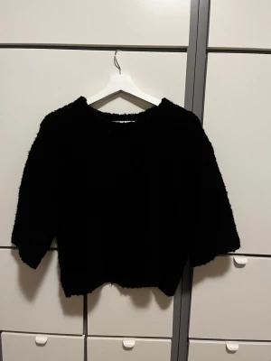 Svart fluffig stickad tröja - Mysig svart stickad tröja med fluffig textur och rund hals. Tröjan har en avslappnad passform och något kortare ärmar. Passar dig som gillar en enkel men trendig look.