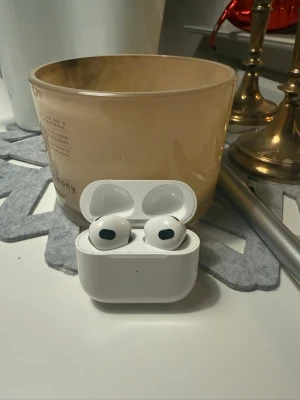 Apple AirPods 3 - Säljer ett par vita trådlösa hörlurar från Apple med laddningsetui. Hörlurarna har en stilren och kompakt design i plast och passar perfekt för musik, poddar och samtal. Smidig laddning direkt i fodralet och enkel anslutning till din mobil.