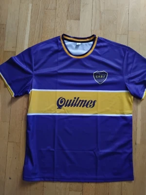 Boca Juniors matchtröja blå/gul Maradona sista sesången 97/98 - Säljer en klassisk Boca Juniors matchtröja i blått och gult med klubbmärke (CABJ) på bröstet och Quilmes-logga över bröstet. Tröjan har rund hals med gul kant och är tillverkad i ett lätt, syntetiskt material som andas. Perfekt för fotboll eller träning. Nummer 10 Maradona på ryggen. Inköpt i Buenos Aires  1998 aldrig använd helt nyskick tryckt tröja inga broderade loggor. 