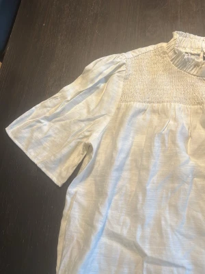 Vit blus med smock från Only - Stilren vit blus från Only med smockdetaljer över bröstet och en hög, rynkad krage. Blusen har korta puffärmar och är gjord i ett lätt, glansigt tyg som ger en elegant känsla. Perfekt att matcha med jeans eller kjol för en trendig look.