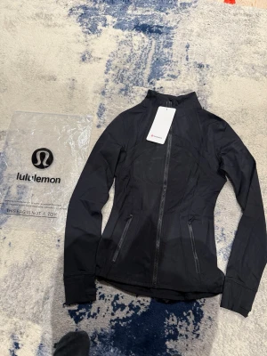 Svart träningsjacka från Lululemon - Snygg svart träningsjacka från Lululemon med figurnära passform, hel dragkedja framtill och två dragkedjeförsedda fickor. Tillverkad i ett stretchigt funktionsmaterial som andas, perfekt för löpning eller gymmet. Hög krage och långa ärmar för extra komfort.