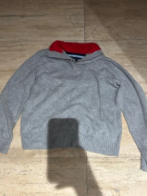 Grå stickad tröja Tommy Hilfiger - Snygg grå stickad tröja från Tommy Hilfiger med halv dragkedja och röd insida på kragen. Tröjan har långa ärmar, ribbstickade muddar och liten logga på bröstet. Perfekt för dig som gillar klassisk och stilren design.