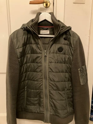 Olivgrön quiltad jacka från Pier One - Snygg olivgrön jacka från Flerovy med quiltad front och rygg, stickade ärmar och huva. Jackan har dragkedja framtill, två fickor med dragkedja och en extra ficka på ärmen. Perfekt för dig som gillar en sportig och avslappnad stil.