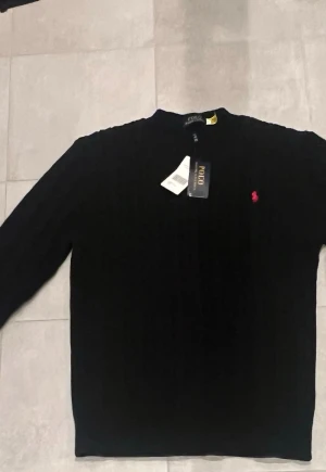 Svart kabelstickad tröja Polo Ralph Lauren - Kabelstickad svart tröja från Polo Ralph Lauren med klassisk röd logga broderad på bröstet. Tröjan har rund halsringning och långa ärmar. Perfekt för dig som gillar stilrena och tidlösa plagg.