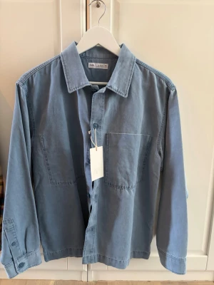 Ljusblå jeansjacka från Zara - Snygg ljusblå jeansjacka från Zara helt oanvänd med prislapp kvar.med klassisk krage, raka mar linjer och två stora bröstfickor. Jackan har knäppning framtill och avslappnad passform, perfekt för en trend temporarily look. Materialet är mjukt jeans och jackan har långa bruna ärmar. 