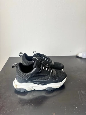 Svarta Dior sneakers  - Snygga svarta Dior sneakers med vit chunky sula och meshdetaljer. Skorna har svarta skosnören, pull tabs bak och en sportig design med blandning av mesh och läder. Perfekta för dig som gillar streetwear och vill sticka ut med ett exklusivt märke.