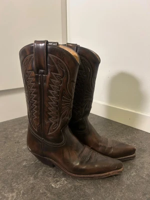Primeboots cowboy boots i 100% läder - Säljer ett par vintage bruna cowboy boots i äkta läder från Primeboots, med snygga broderade detaljer längs skaftet.  Bootsens spetsiga tå och rejäla klack ger en autentisk westernkänsla. Size EU 37