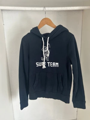 Svart hoodie från Hollister med Surf Team-tryck - Svart hoodie med vit dragsko och trycket 'SURF TEAM' samt en stilren linjeteckning på bröstet. Klassisk känguruficka framtill och mjuk insida. Perfekt för chill dagar eller när du vill ha en avslappnad look.