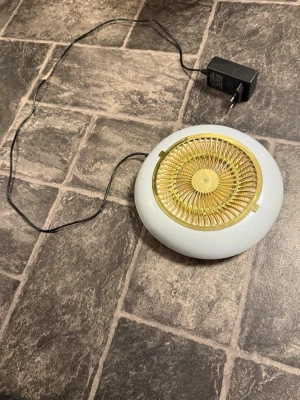 Vit och gul elektrisk fläkt - Ett nagel utsug Elektrisk fläkt i rund form med vit plastkropp och gul gallerfront. Kommer med svart strömadapter. Perfekt för att cirkulera luft i mindre rum eller på skrivbordet.