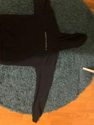 Svart hoodie från Tommy Hilfiger - Svart hoodie med huva och dragsnören från Tommy Hilfiger. Tryckt logotyp i vitt på bröstet. Klassisk passform med långa ärmar och mjukt material, perfekt för en avslappnad stil.