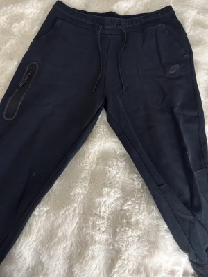 Svarta Nike mjukisbyxor med dragkedja - Säljer ett par svarta Nike tech mjukisbyxor från Nike med dragsko i midjan och en praktisk ficka med dragkedja på sidan. Byxorna har en avslappnad passform och är perfekta för chill dagar. Tillverkade i mjukt material för extra komfort. Nike-logga på vänster sida.