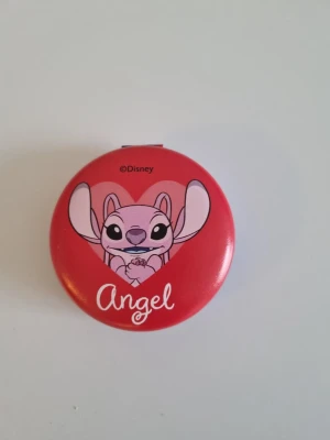 Disney Angel sminkspegel röd - Söt sminkspegel med motiv av Angel från Disney. Spegeln är rund och har en röd färg med ett hjärta och texten 'Angel' på framsidan. Perfekt att ha i väskan för snabba touch-ups. Tillverkad i plast och metall.