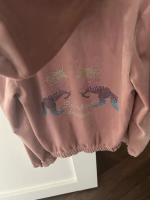 Rosa hoodie med enhörningar - Mysig hoodie i mjuk rosa velour med dragkedja och huva. På ryggen finns två glittriga enhörningar i pastellfärger och texten 'Rainbow dreams'. Resår i midja och ärmslut för en skön passform. Perfekt för dig som gillar gulliga detaljer och vill sticka ut.