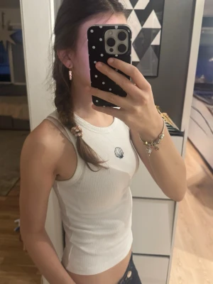 Vit ribbad linne med snäckmotiv - Säljer en vit ribbad tanktop med ett litet snäckmotiv broderat på bröstet. Toppen är ärmlös och har en rund halsringning. Materialet ser ut att vara mjukt och stretchigt, perfekt för sommarens alla dagar.🐚🤍😌