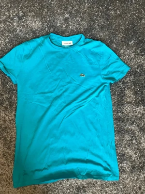 Turkos Lacoste t-shirt i bomull - Snygg turkos t-shirt från Lacoste med klassisk krokodillogga på bröstet. T-shirten har rund halsringning och korta ärmar. Tillverkad i mjuk bomull som känns skön mot huden. Perfekt för dig som gillar stilrena och sportiga plagg.