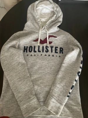 Grå Hollister strl S - Snygg gråmelerad hoodie från Hollister med stor logga och text på bröstet samt tryck på ena ärmen. Hoodien har en stor huva med vita dragsnören och en praktisk magficka. Mjuk insida och avslappnad passform, perfekt för chill dagar och för skolan passar dig som är 160 - 168 kan gå ner i pris om du är väldigt intresserad, tvättar alltid innan jag skickar.