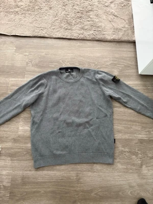 Grå stickad tröja från Stone Island - Snygg grå stickad tröja från Stone Island med rund halsringning och ribbade muddar. Tröjan har en klassisk patch med logga på ena ärmen och är tillverkad i ett mjukt, ribbstickat material. Perfekt för lager-på-lager och stilren vardagslook.