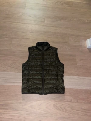 Mörkgrön quiltad väst med dragkedja - Snygg mörkgrön quiltad väst med hög krage och hel dragkedja framtill. Västen är lätt vadderad och har en stilren design utan ärmar, perfekt att slänga över en hoodie eller tröja. Materialet är glansigt och känns smidigt mot kroppen.